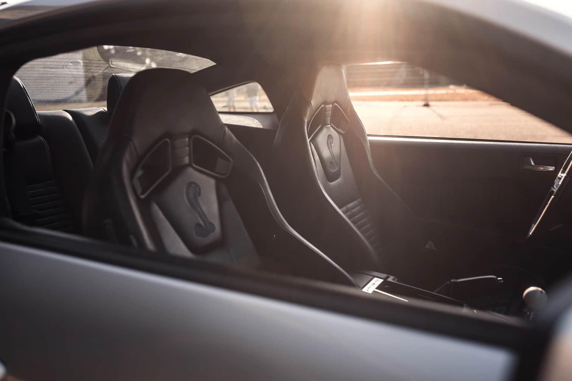 Detail des dunklen Sportwagen-Innenraums mit Schalensitzen und Cobra-Logo, Gegenlicht der tiefstehenden Sonne beim Automotive-Shooting