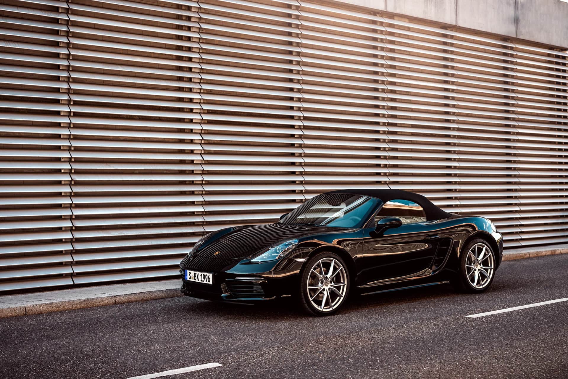 Schwarzer Porsche 718 Boxster schraeg von vorn vor einer horizontal gegliederten Metallfassade, glaenzender Lack im warmen Car-Shooting-Licht