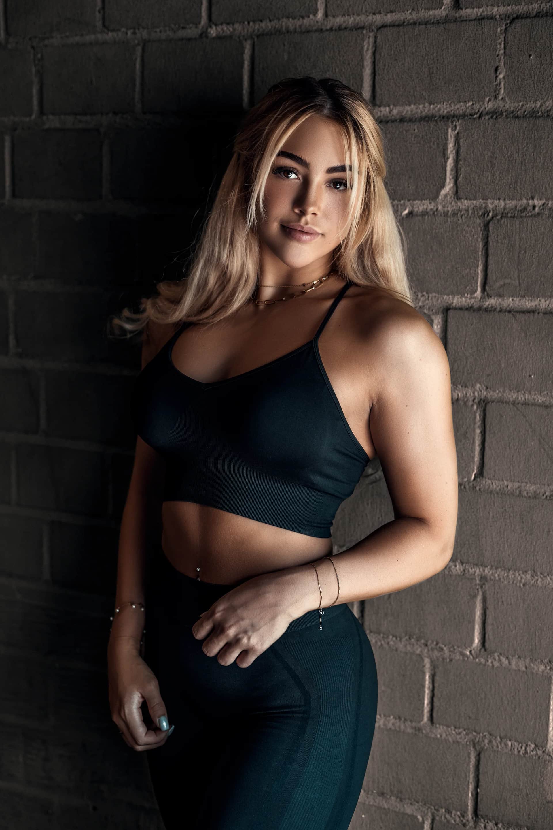 Blonde Frau im schwarzen Sport-Outfit lehnt an einer dunklen Backsteinwand, durchtrainierter Look beim Fitness-Shooting in stimmungsvollem Licht