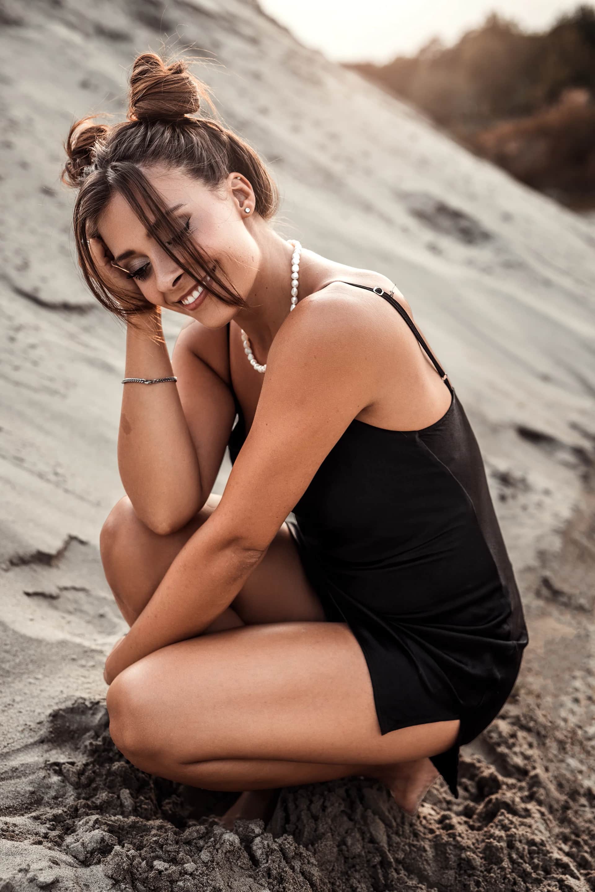 Junge Frau im schwarzen Trägertop mit Perlenkette kauert lachend in einer Sanddüne, warmes Gegenlicht im Abendlicht – natürliches Portraitshooting