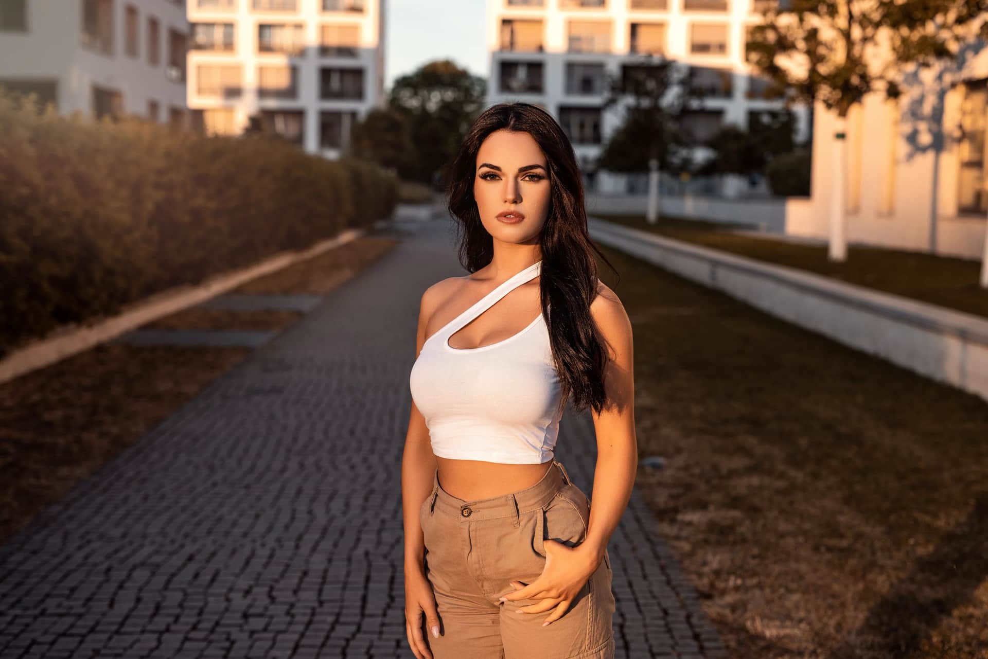 Portraitshooting im warmen Abendlicht: dunkelhaarige Frau im weissen One-Shoulder-Top und beiger Hose auf gepflastertem Weg vor moderner Wohnbebauung.