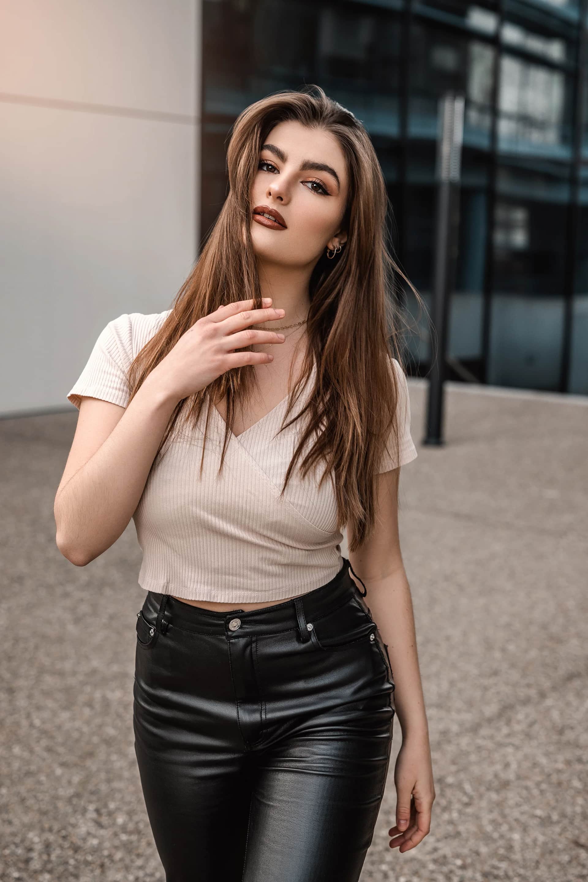 Frau in beigem Crop-Top und schwarzer Lederhose posiert vor moderner Glasfassade, lange Haare im Wind – urbanes Portraitshooting