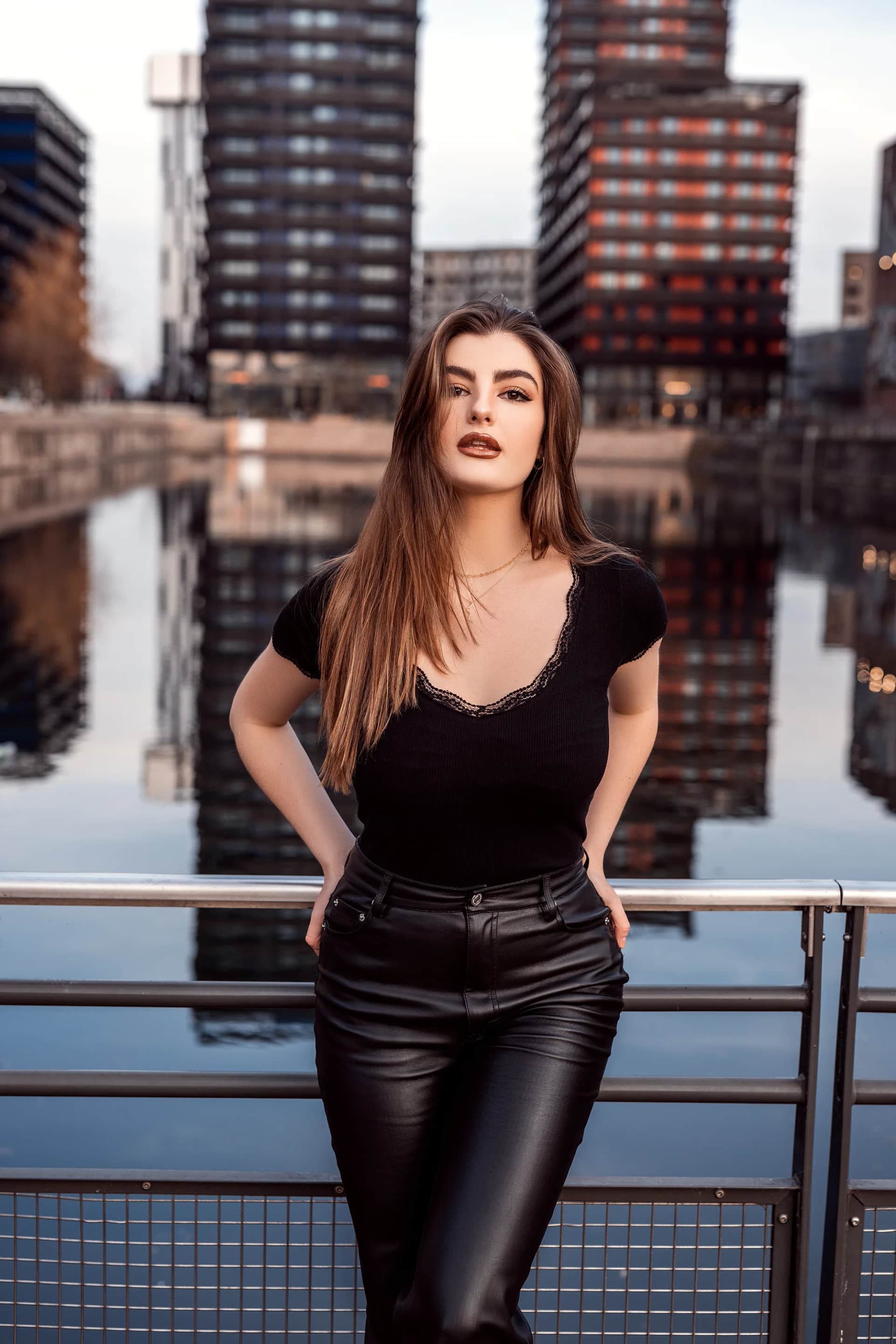 Frau mit langen Haaren in schwarzem Top und Lederhose lehnt an einem Geländer vor Hochhäusern, die sich im Wasser spiegeln – urbanes Portraitshooting