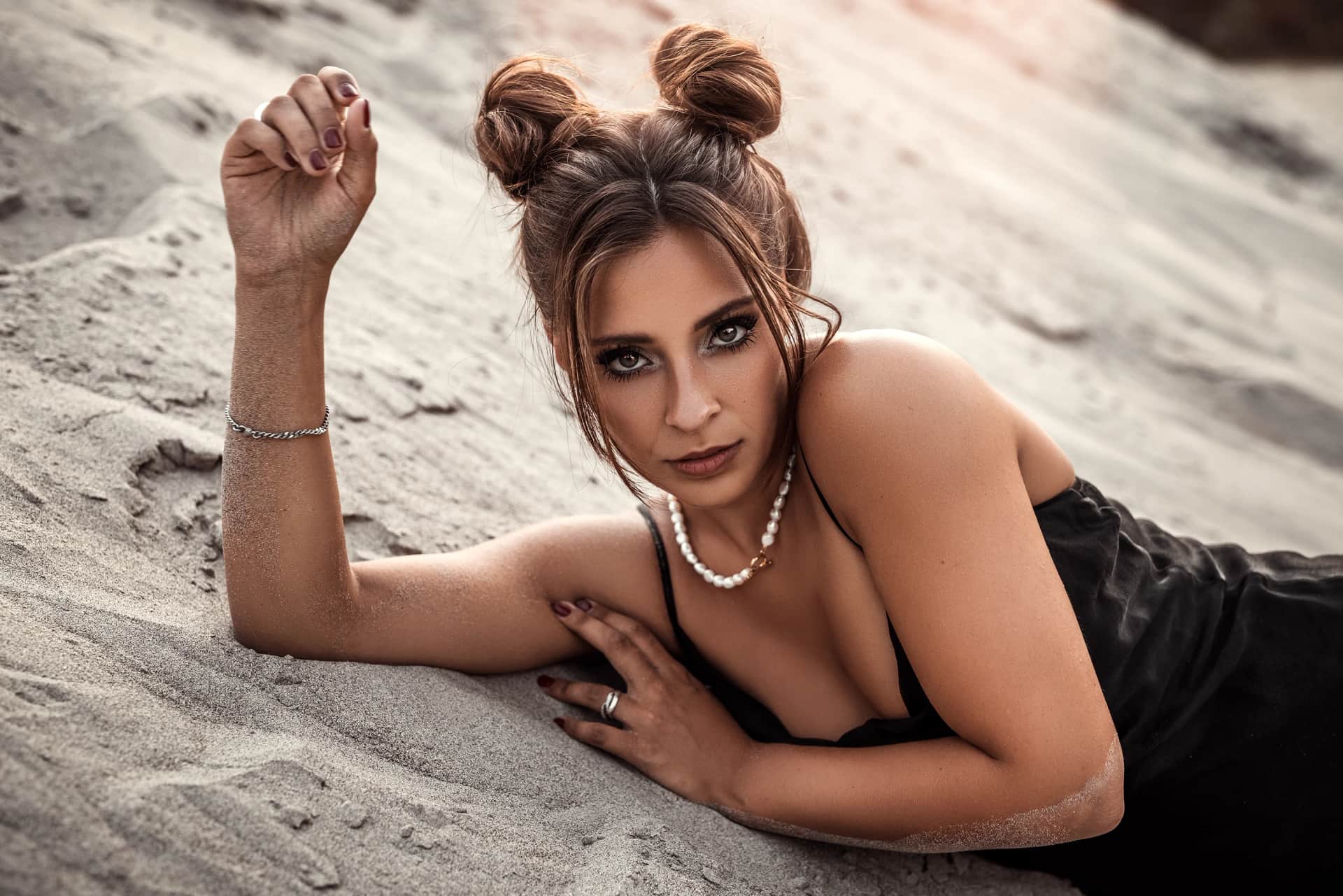Portraitshooting im Sand: Frau mit Space-Buns, Perlenkette und schwarzem Kleid liegt elegant auf einer Duene, intensiver Blick in die Kamera.
