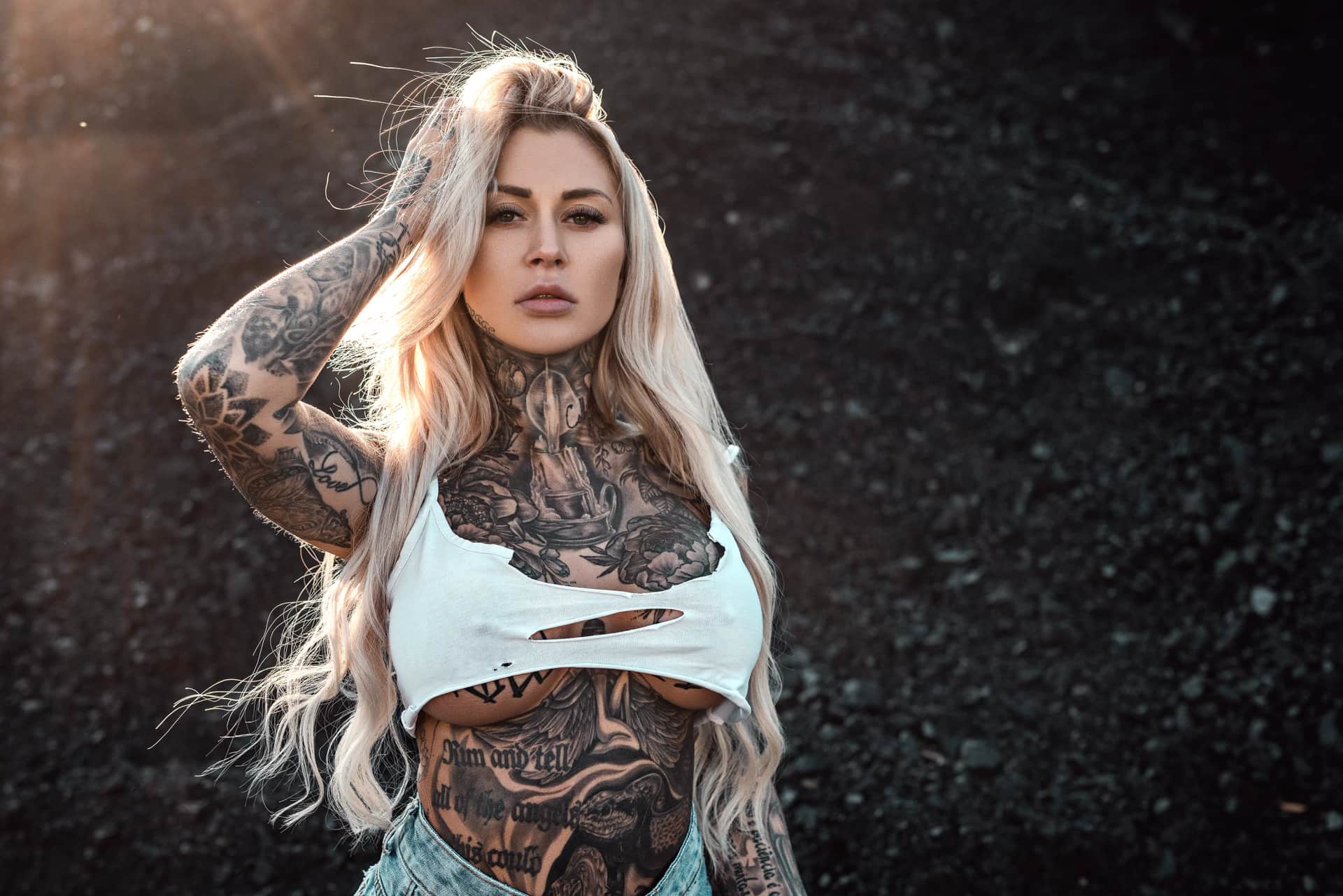 Portraitshooting einer blonden, taetowierten Frau im weissen Crop-Top vor dunkler Kieskulisse, Hand im Haar, intensiver Blick im Gegenlicht.