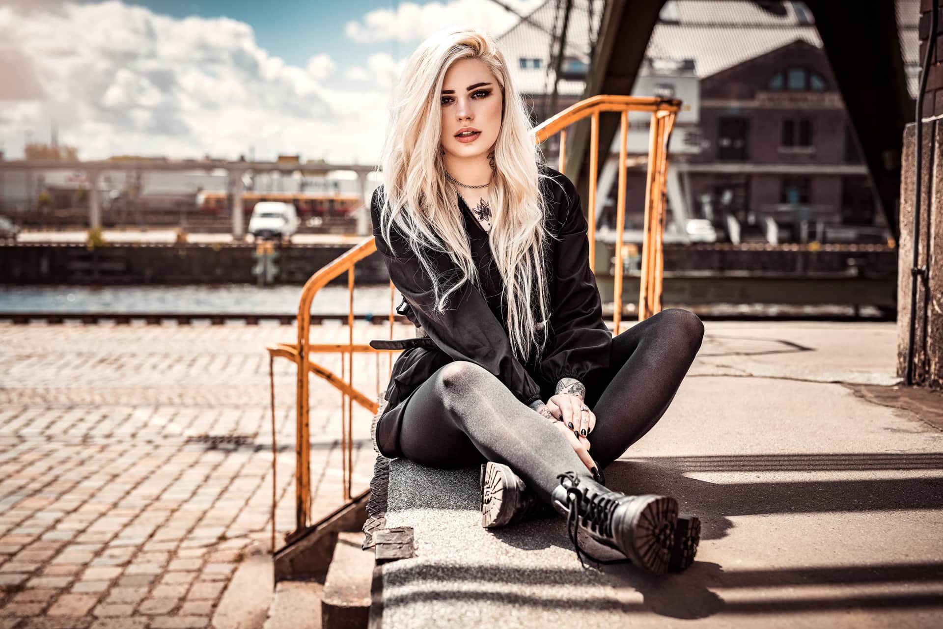 Blonde Frau in Lederjacke und Boots sitzt lässig auf einer Kaimauer im Industriehafen, Portraitshooting mit Backsteingebäuden und blauem Himmel