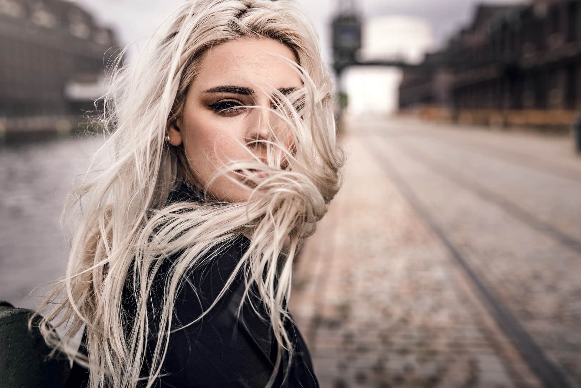 Blonde Frau mit windverwehten Haaren und Rauchmake-up blickt intensiv in die Kamera, urbanes Portraitshooting an grauer Stadtkulisse