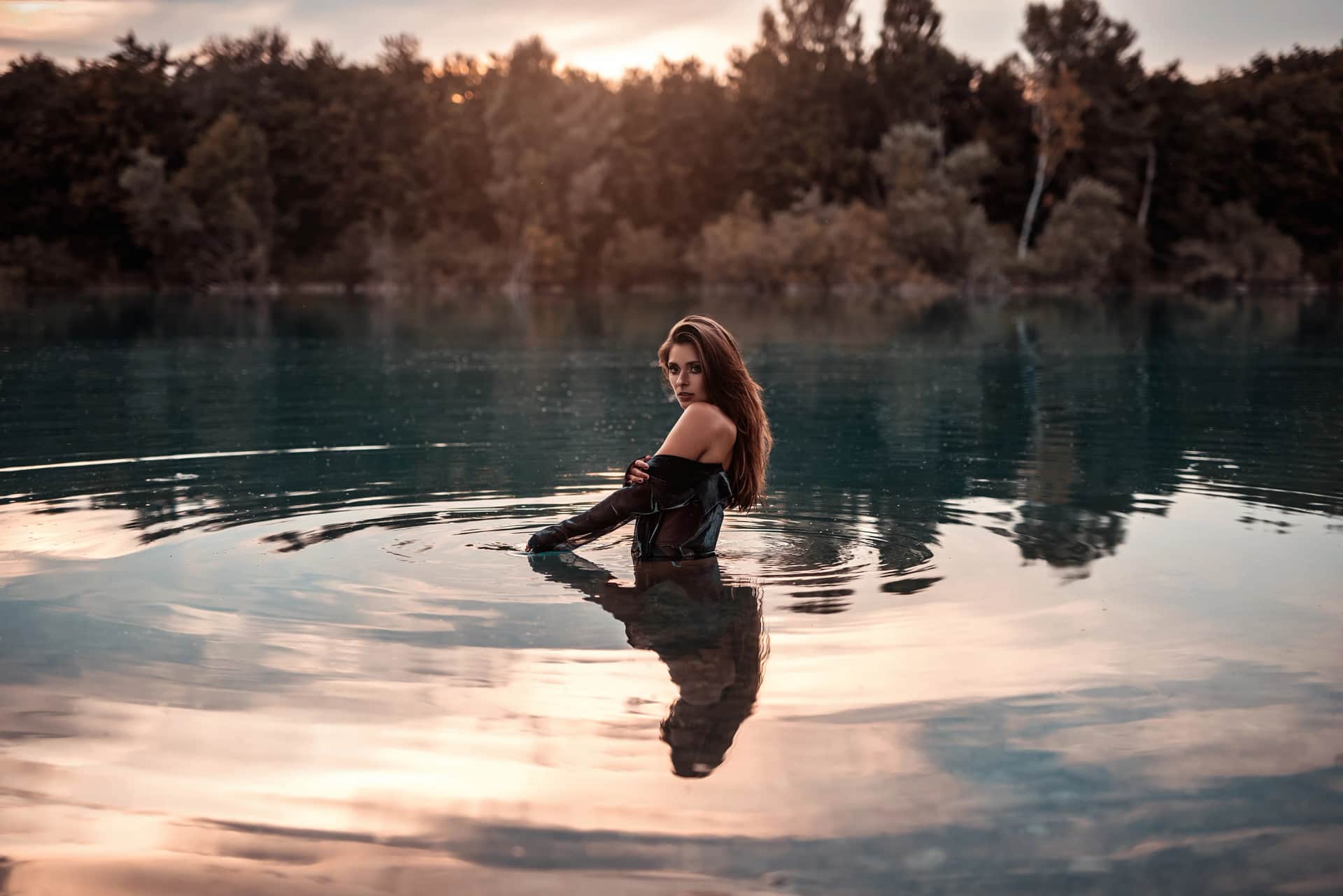 Stimmungsvolles Portraitshooting: Frau in dunkler Kleidung steht huefttief in einem ruhigen See, gespiegelt im Wasser, bewaldetes Ufer im Sonnenuntergang.