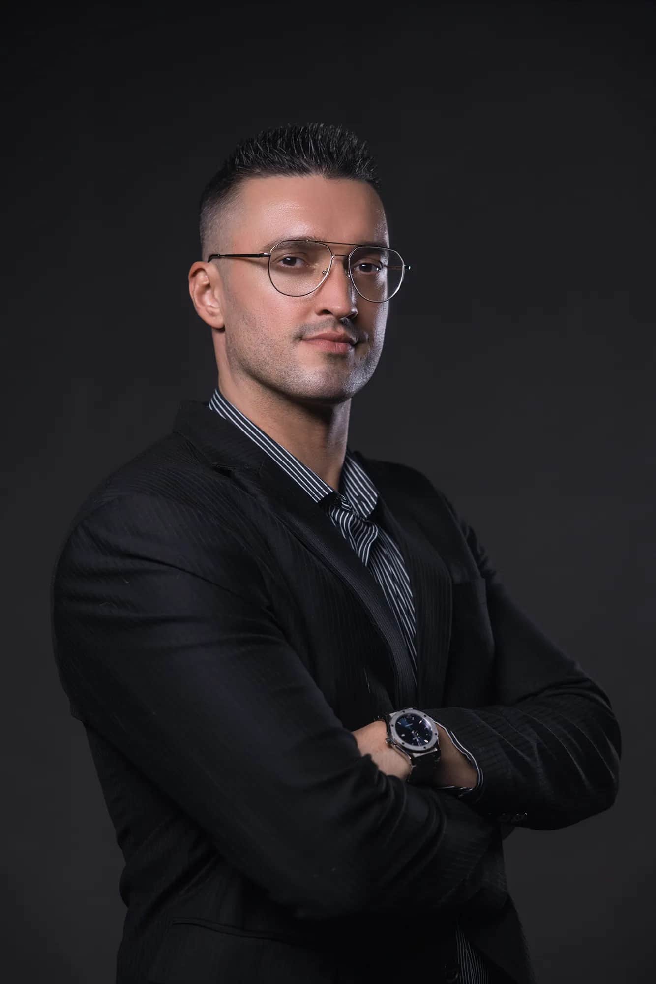 Mann mit Brille und Dreitagebart in schwarzem Sakko und gestreiftem Hemd, verschraenkte Arme, markantes Business-Portrait vor dunklem Studiohintergrund.