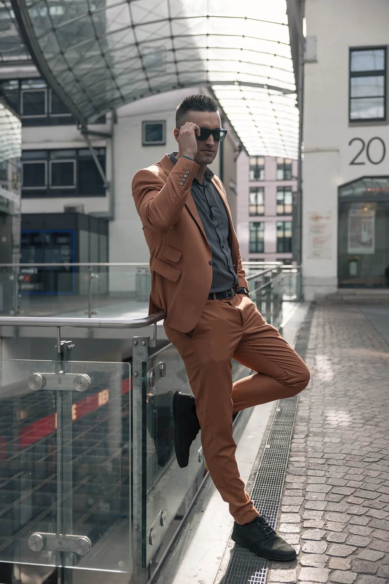 Mann im rostbraunen Anzug mit Sonnenbrille lehnt lässig an einem Geländer unter einem Glasdach, urbanes Business-Portrait mit kühlem Tageslicht