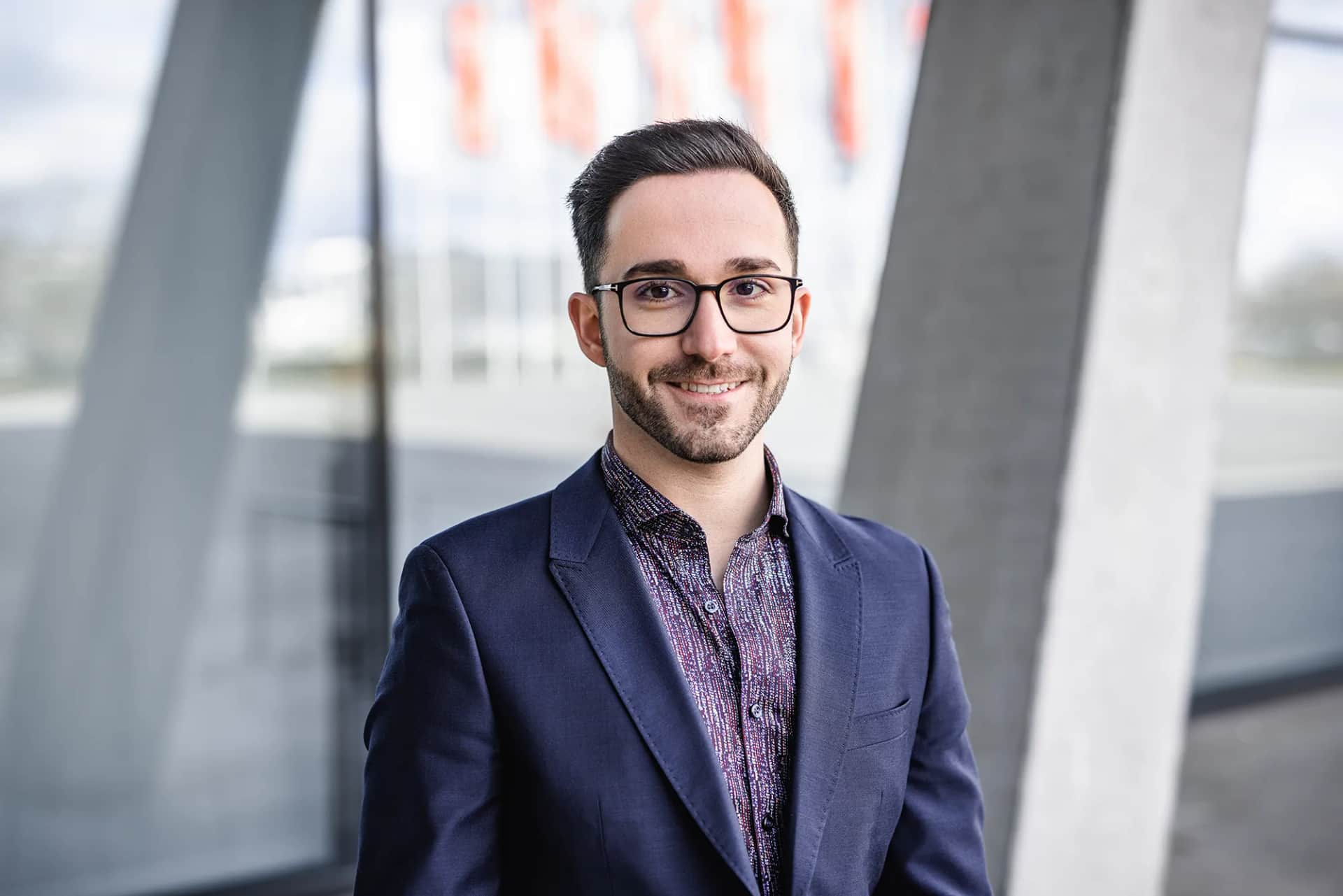 Laechelnder Mann mit Brille im dunkelblauen Sakko vor moderner Architektur, freundliches Business-Portrait bei Tageslicht.