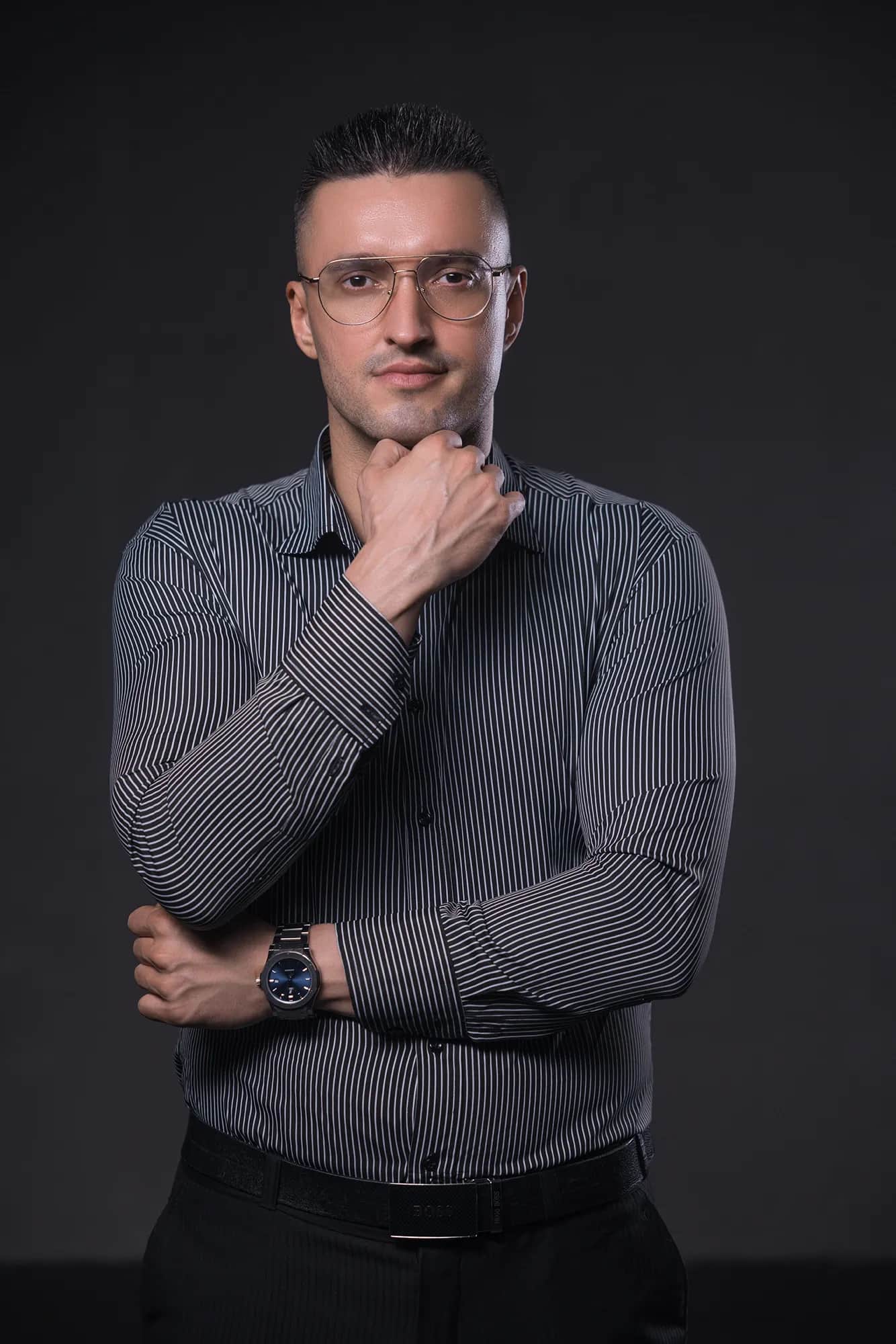 Mann mit Brille in gestreiftem Hemd stuetzt nachdenklich das Kinn, elegantes Business-Portrait vor dunklem Studiohintergrund mit gerichtetem Licht.