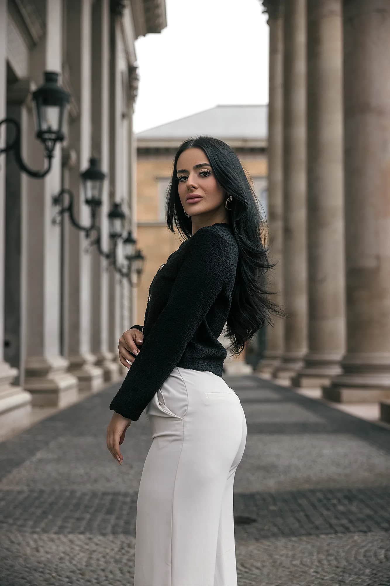 Dunkelhaarige Frau in schwarzem Twiststrick-Top und weisser Hose blickt ueber die Schulter, elegantes Business-Portrait zwischen klassischen Saeulen einer Kolonnade.