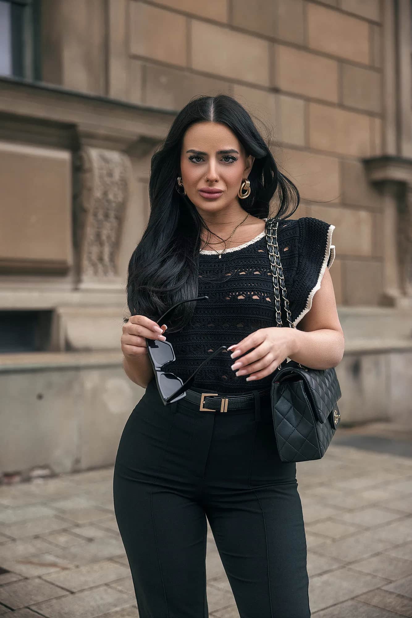 Dunkelhaarige Frau in schwarzem Outfit mit Sonnenbrille und gesteppter Kettentasche vor heller Altbaufassade, urbanes Business-Portrait in gedaempftem Licht.