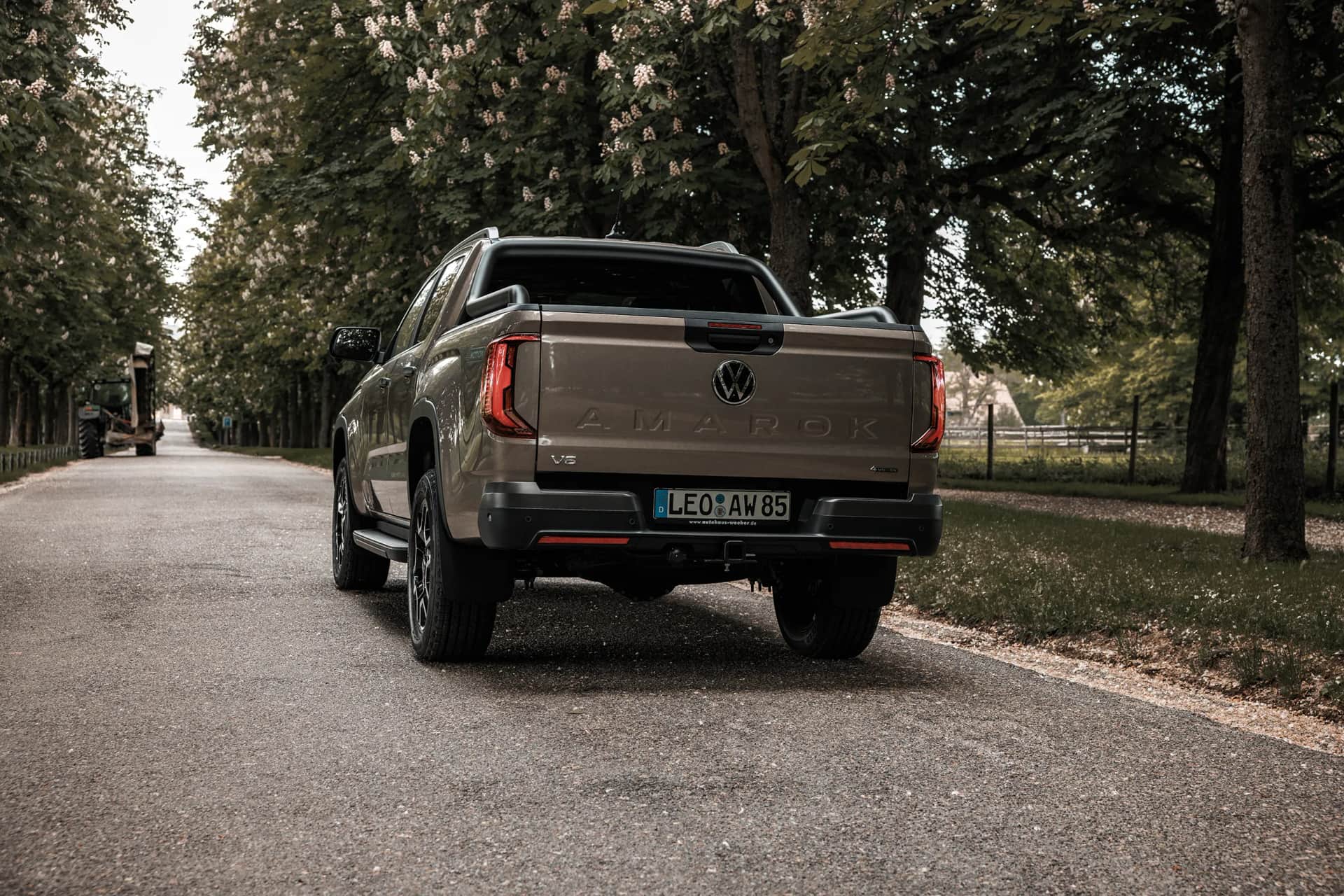 VW Amarok Pickup von hinten auf ruhiger Baumallee, bronzefarbener Lack im weichen Sonnenlicht, Automotive-Shooting