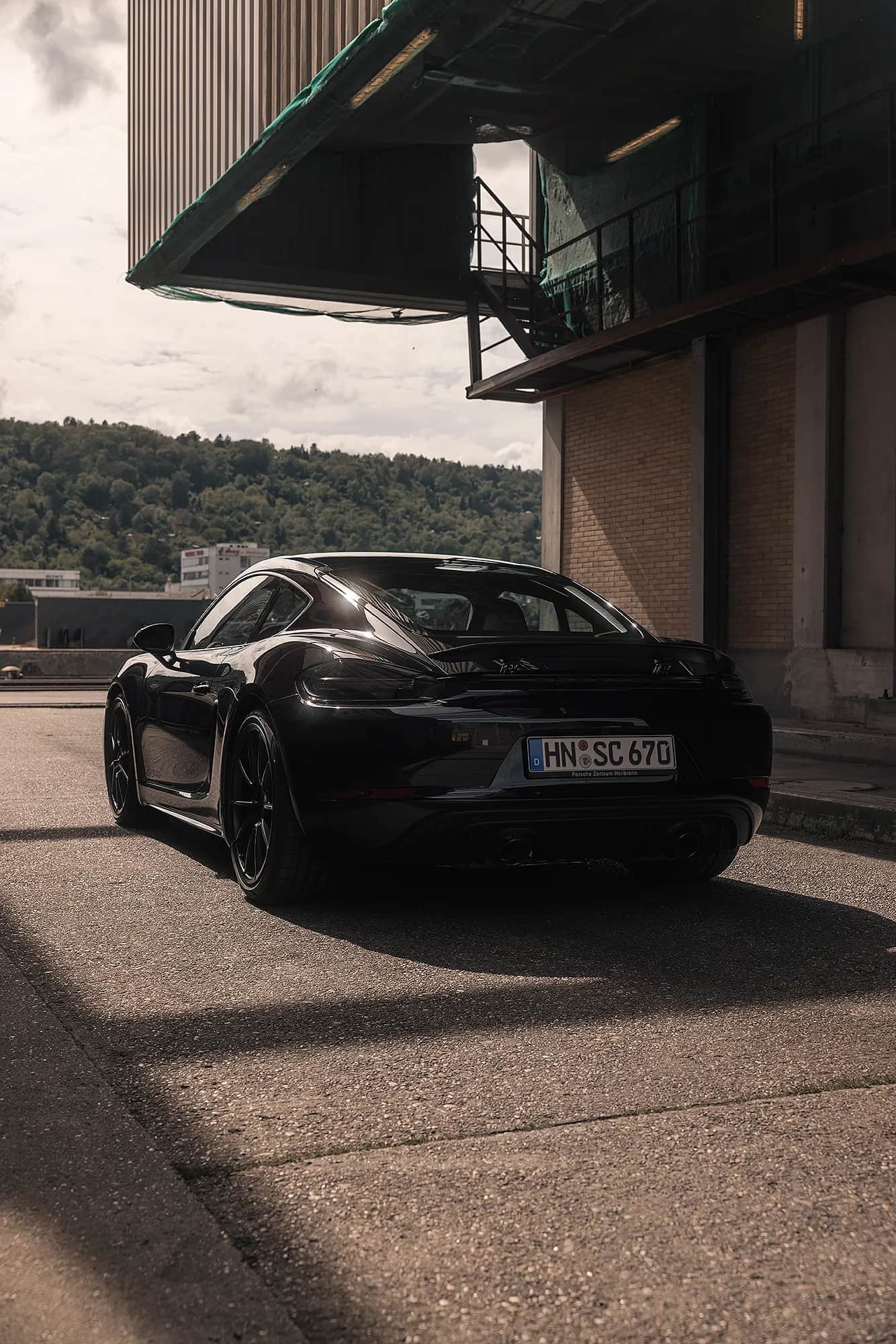 Schwarzer Porsche Coupe von hinten vor Industriehalle mit Blick auf bewaldete Huegel, stimmungsvolles Automotive-Shooting