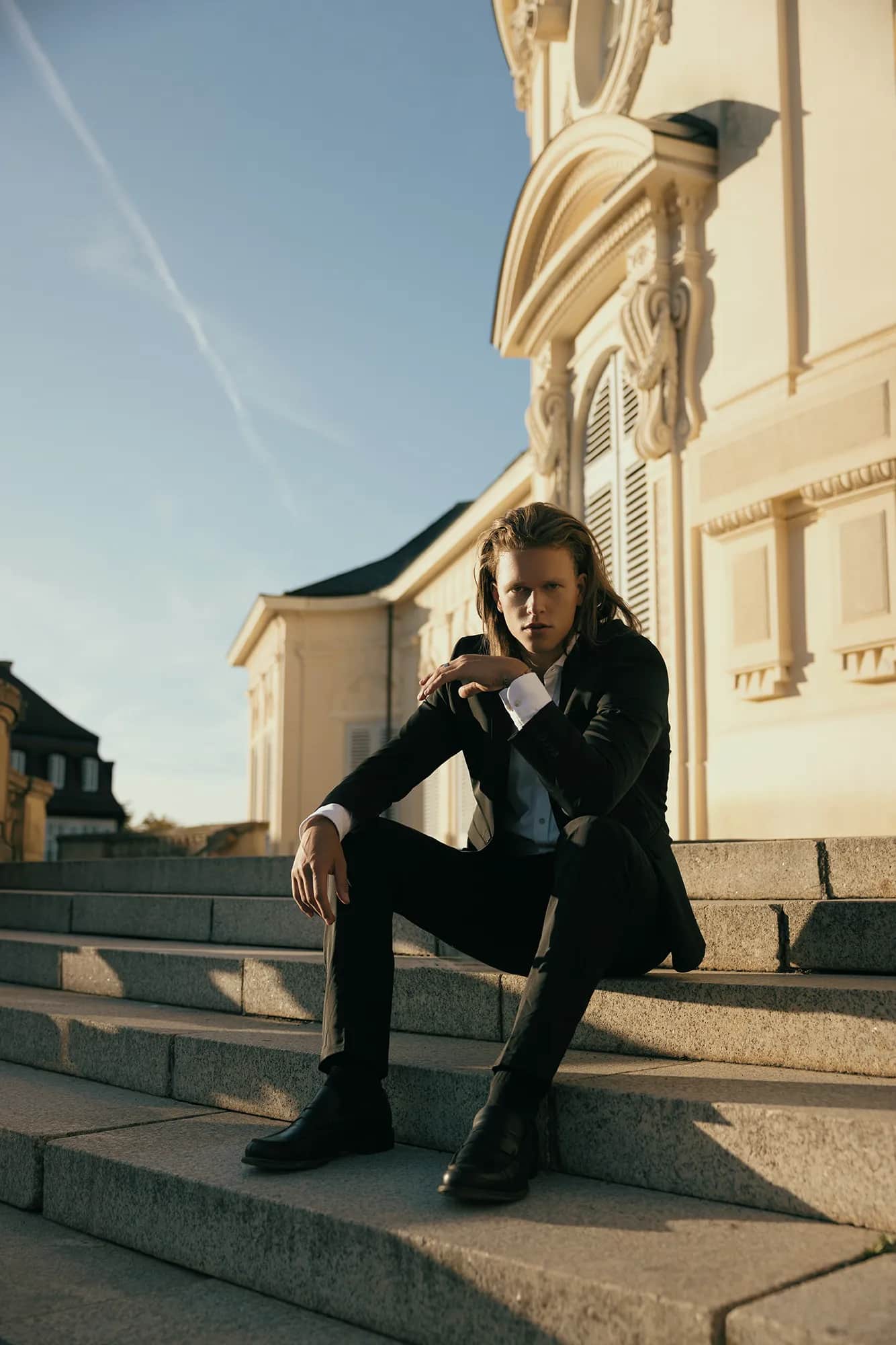 Langhaariger Mann im schwarzen Anzug sitzt auf der Treppe vor barocker Schlossfassade, elegantes Editorial-Shooting im goldenen Abendlicht
