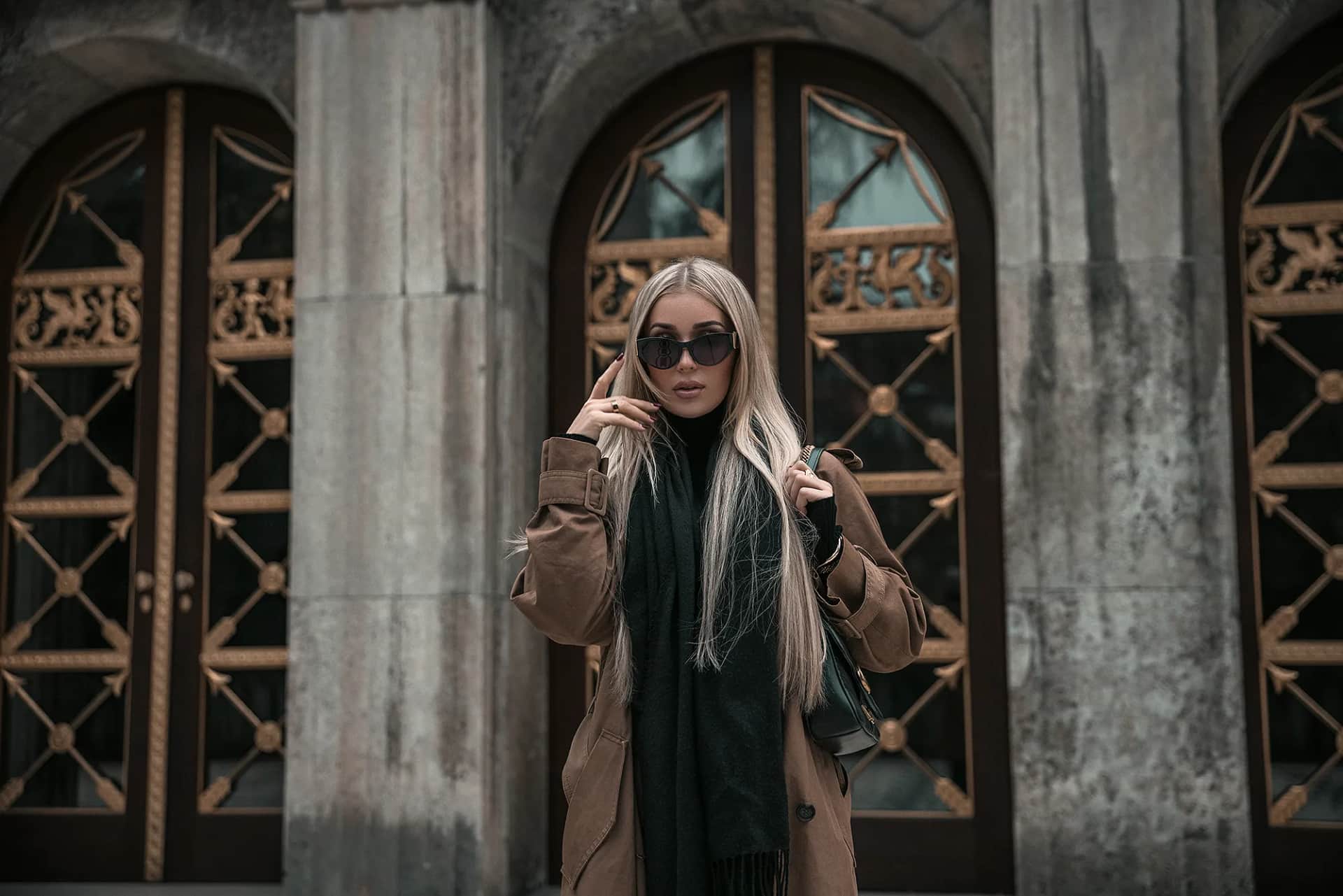 Blondes Model im Camel-Mantel mit Sonnenbrille vor verzierten Bogentueren, gedaempfte Stadtkulisse, urbanes Fashion-Editorial