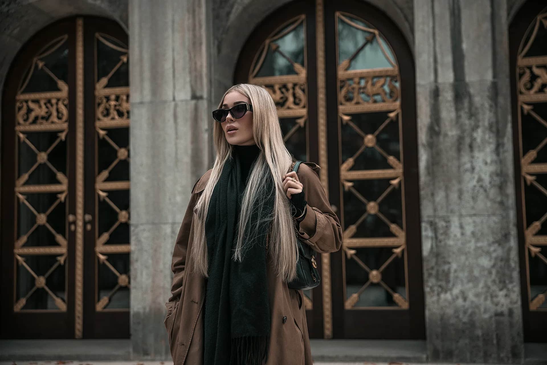 Blonde Frau im Camel-Mantel und mit Sonnenbrille vor schmiedeeisernen Bogenportalen, herbstliche Stimmung, urbanes Fashion-Shooting