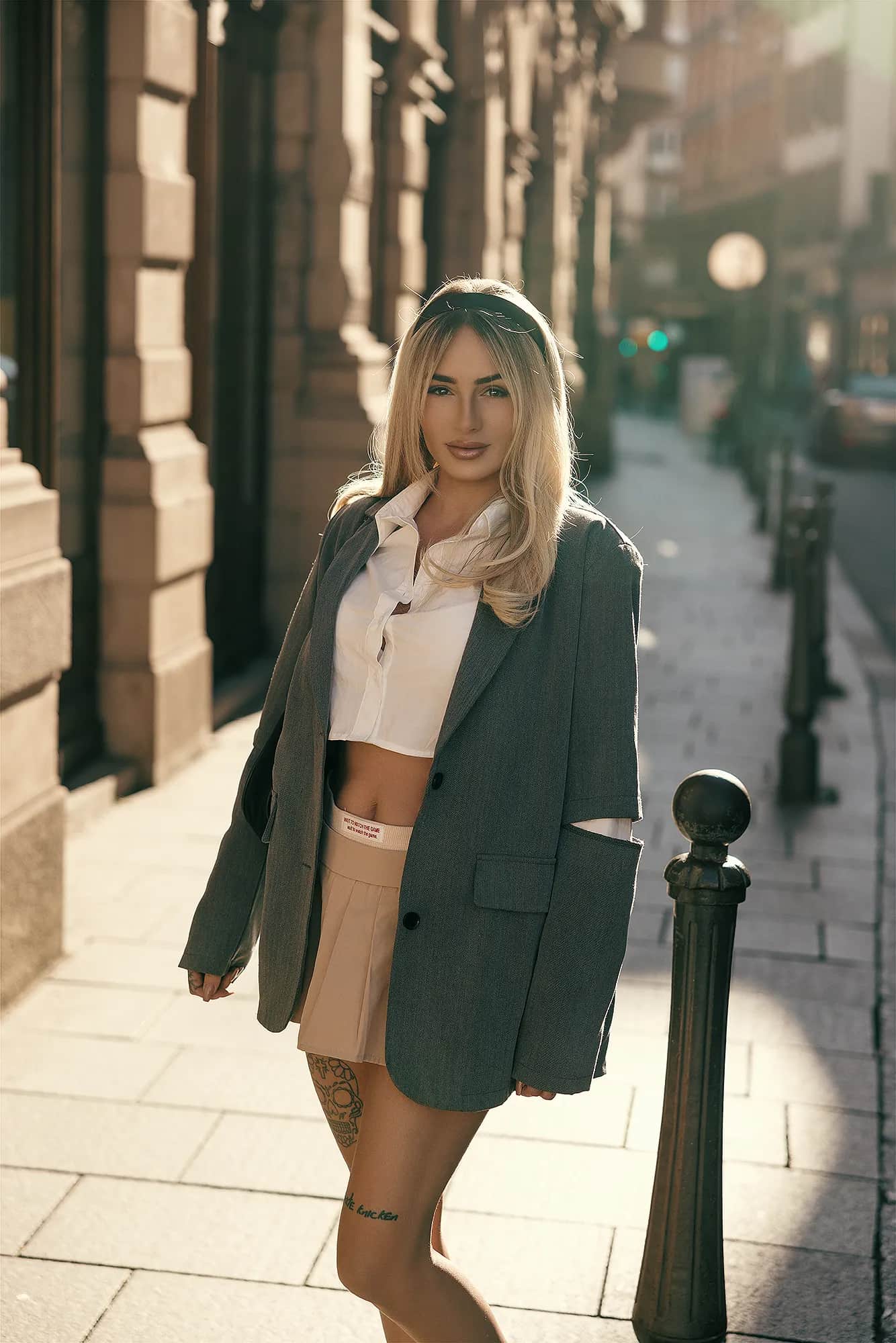 Blonde Frau im grauen Oversize-Blazer ueber weissem Crop-Top und beigem Faltenrock in einer sonnendurchfluteten Altstadtgasse, urbanes Fashion-Editorial
