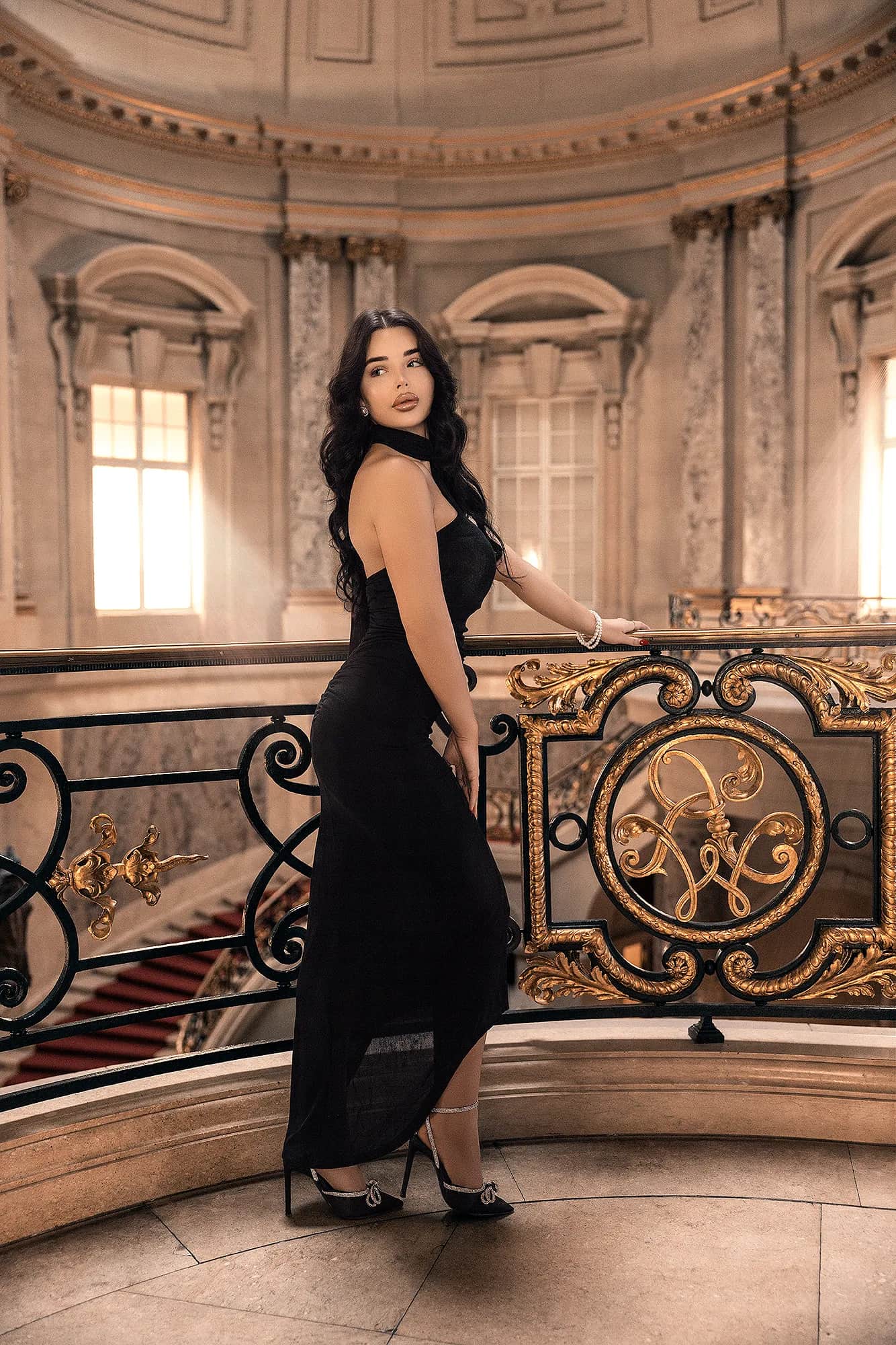 Dunkelhaarige Frau im eleganten schwarzen Abendkleid an einer goldverzierten Balustrade in prunkvollem Treppenhaus, glamouroeses Fashion-Shooting