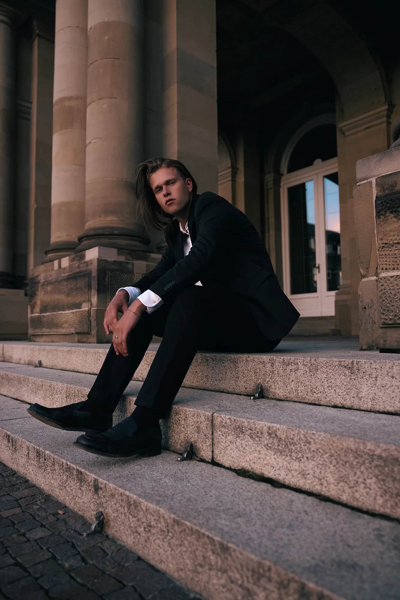 Langhaariger Mann im schwarzen Anzug sitzt auf Steinstufen zwischen Sandsteinsäulen, stimmungsvolles Editorial-Shooting in der Dämmerung