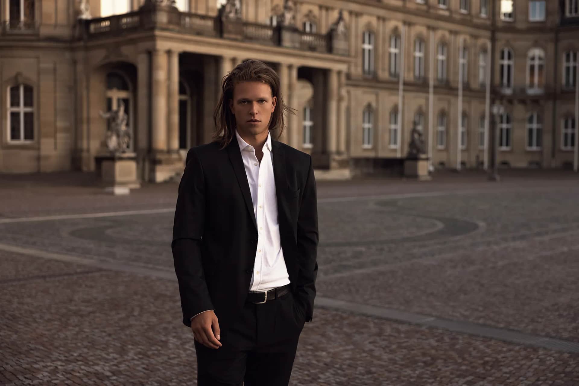 Maennliches Model in schwarzem Anzug mit weissem Hemd steht auf dem kopfsteingepflasterten Schlossplatz vor dem Neuen Schloss Stuttgart, Editorial-Fashion-Shooting bei Sonnenuntergang