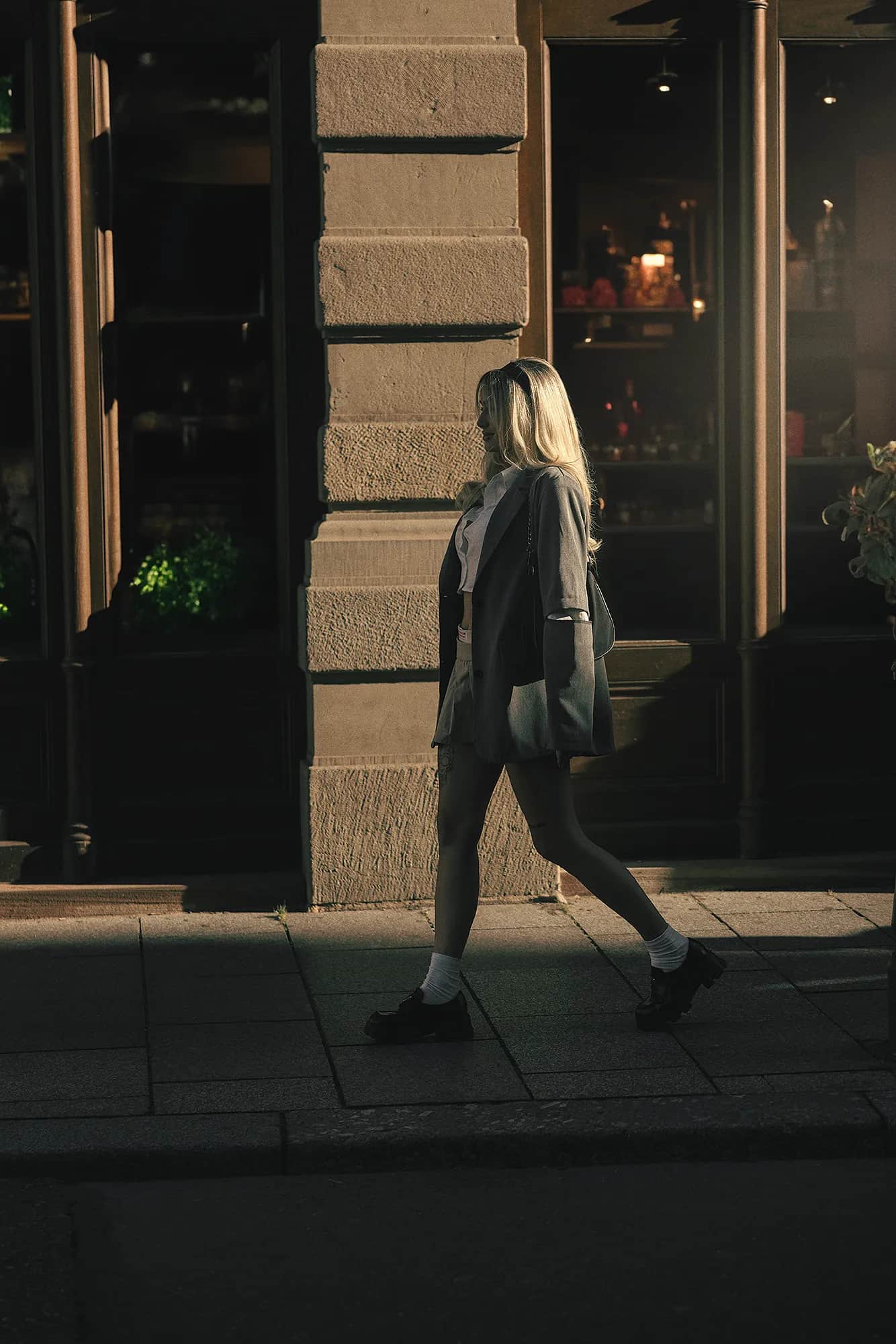 Blonde Frau im grauen Oversize-Blazer geht abends an einer beleuchteten Schaufensterfront entlang, stimmungsvolles urbanes Fashion-Shooting in der Daemmerung
