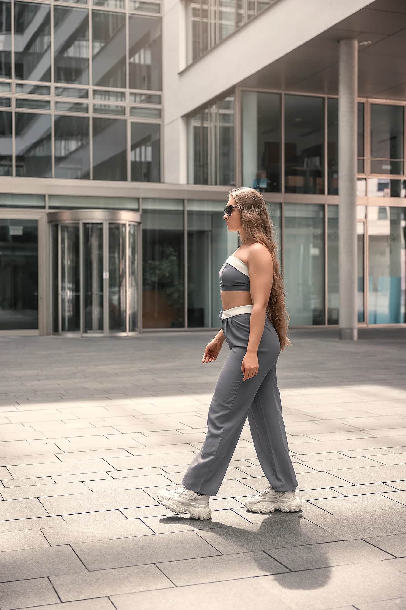 Junge Frau mit langem blondem Haar und Sonnenbrille im grauen Two-Piece schreitet vor verglaster Bueroarchitektur entlang, urbanes Business-Shooting in Tageslicht.