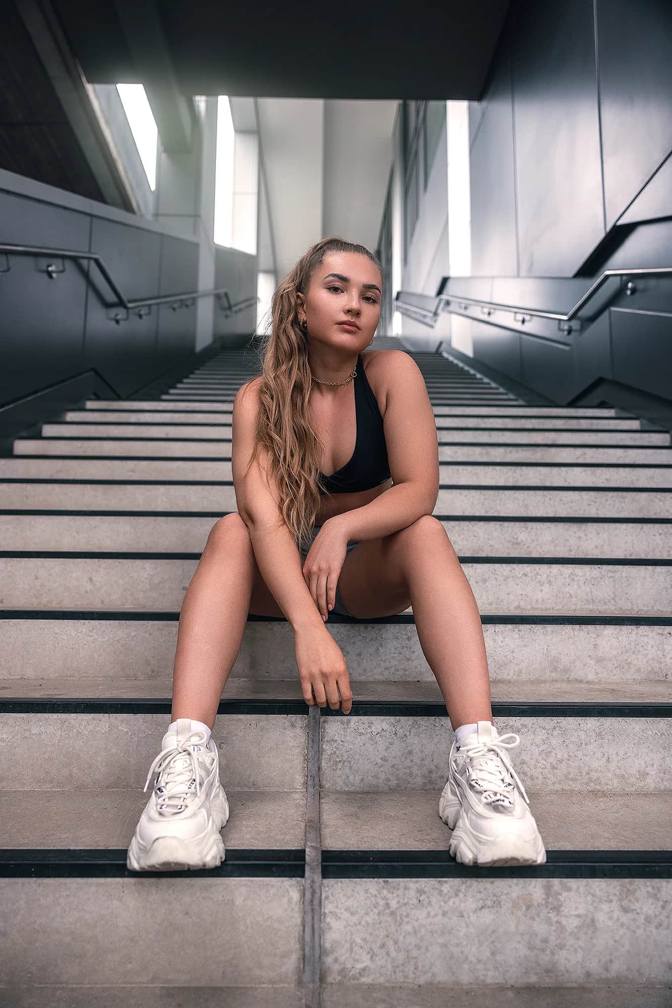 Junge Frau im schwarzen Sport-Bra sitzt auf einer modernen Betontreppe in urbaner Architektur, sportlich-cooles Fitness-Shooting bei kühlem Tageslicht