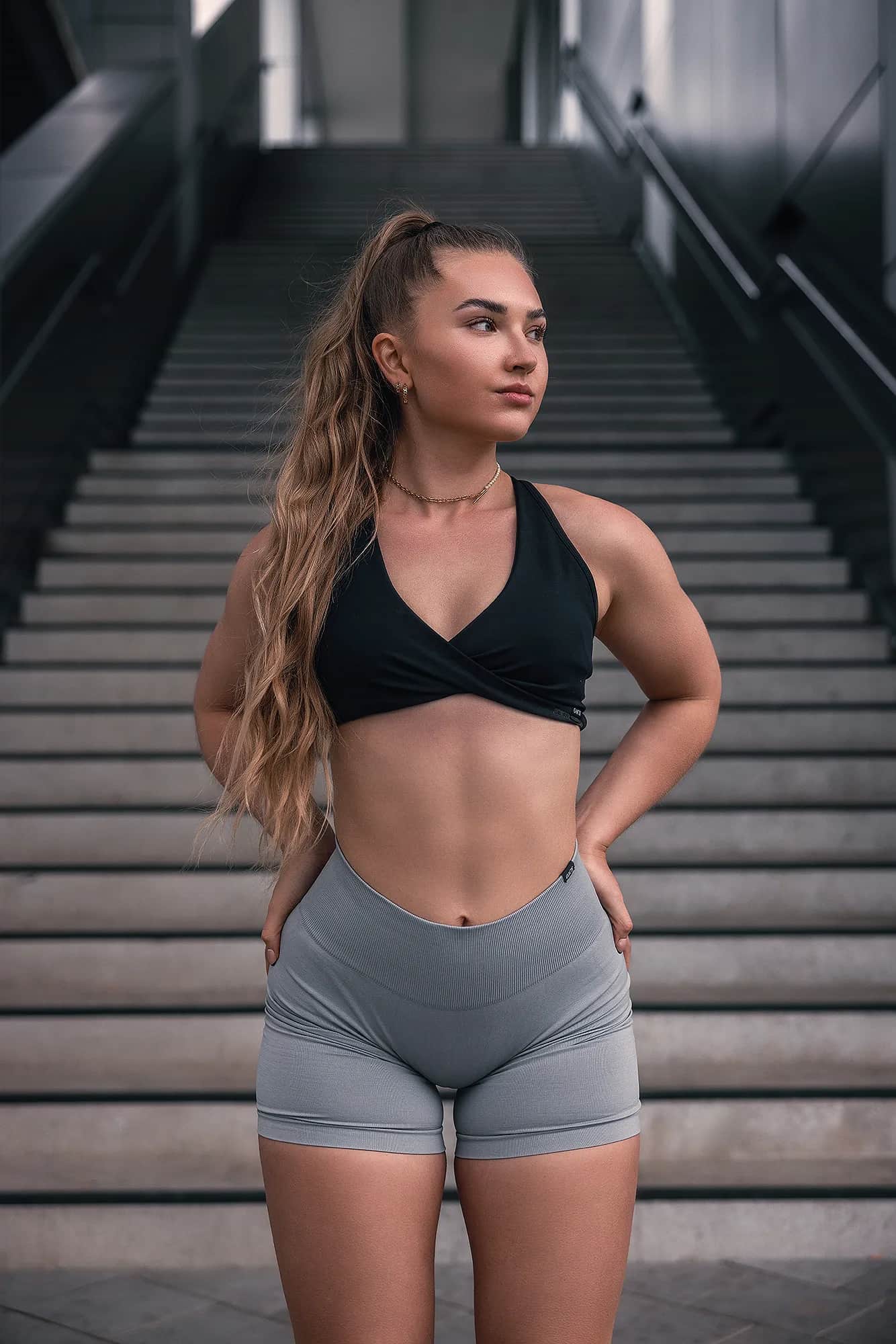 Sportlerin in schwarzem Sport-BH und grauer Shorts steht vor einer Betontreppe, definierte Bauchmuskeln – urbanes Fitness-Shooting