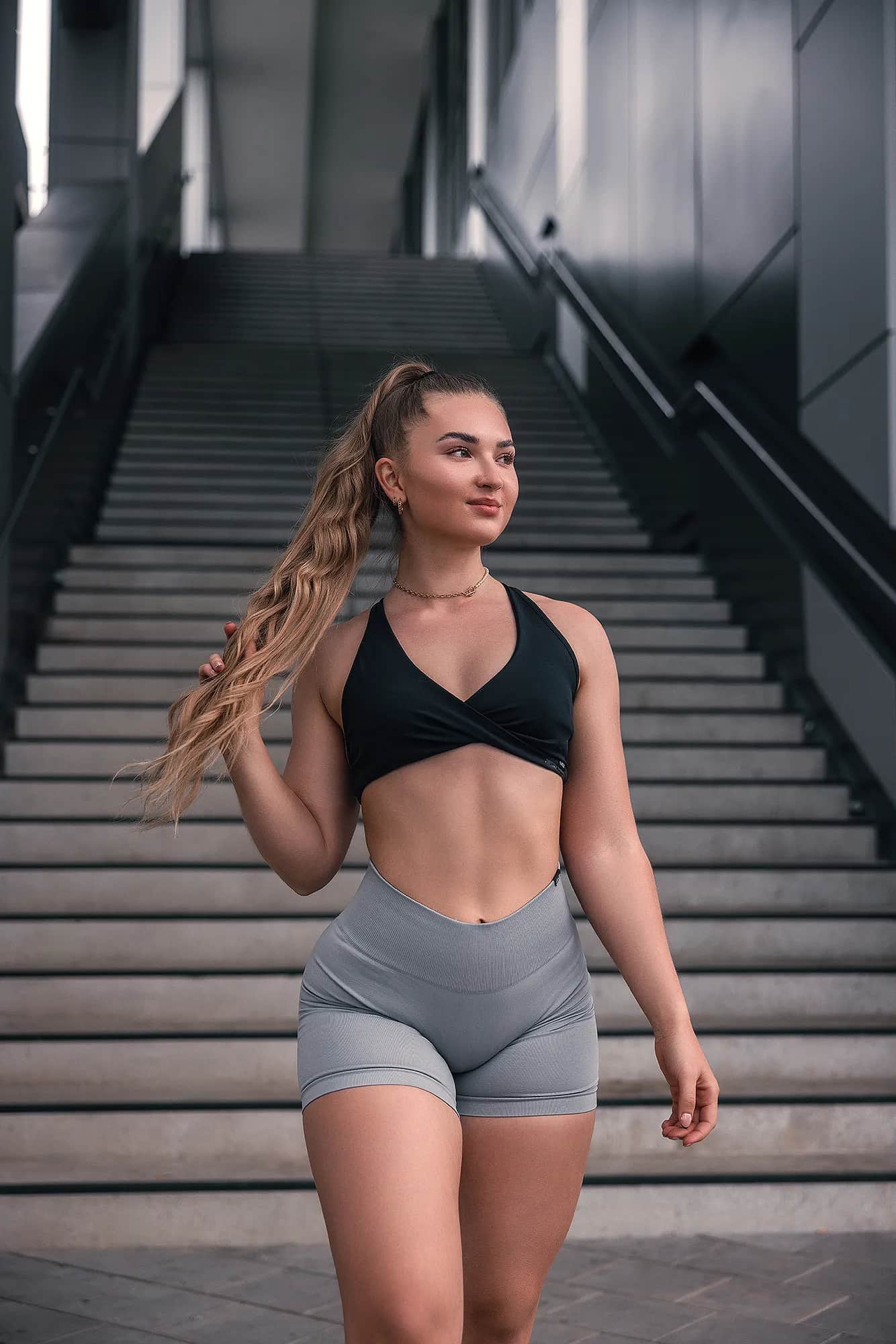 Lächelnde Athletin in Sportoutfit greift in ihren langen Pferdeschwanz vor breiter Außentreppe – dynamisches Fitness-Shooting