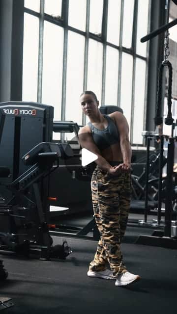 Muskulöse Person in Camouflage-Hose und Crop-Top beim Training im Gym, dramatisches Gegenlicht durch große Fenster