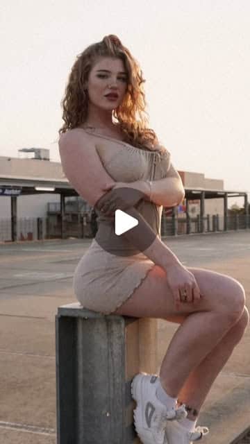 Junge Frau mit roten Locken im beigen Sommeroutfit auf einem Pfosten sitzend, urbanes Editorial im Abendlicht