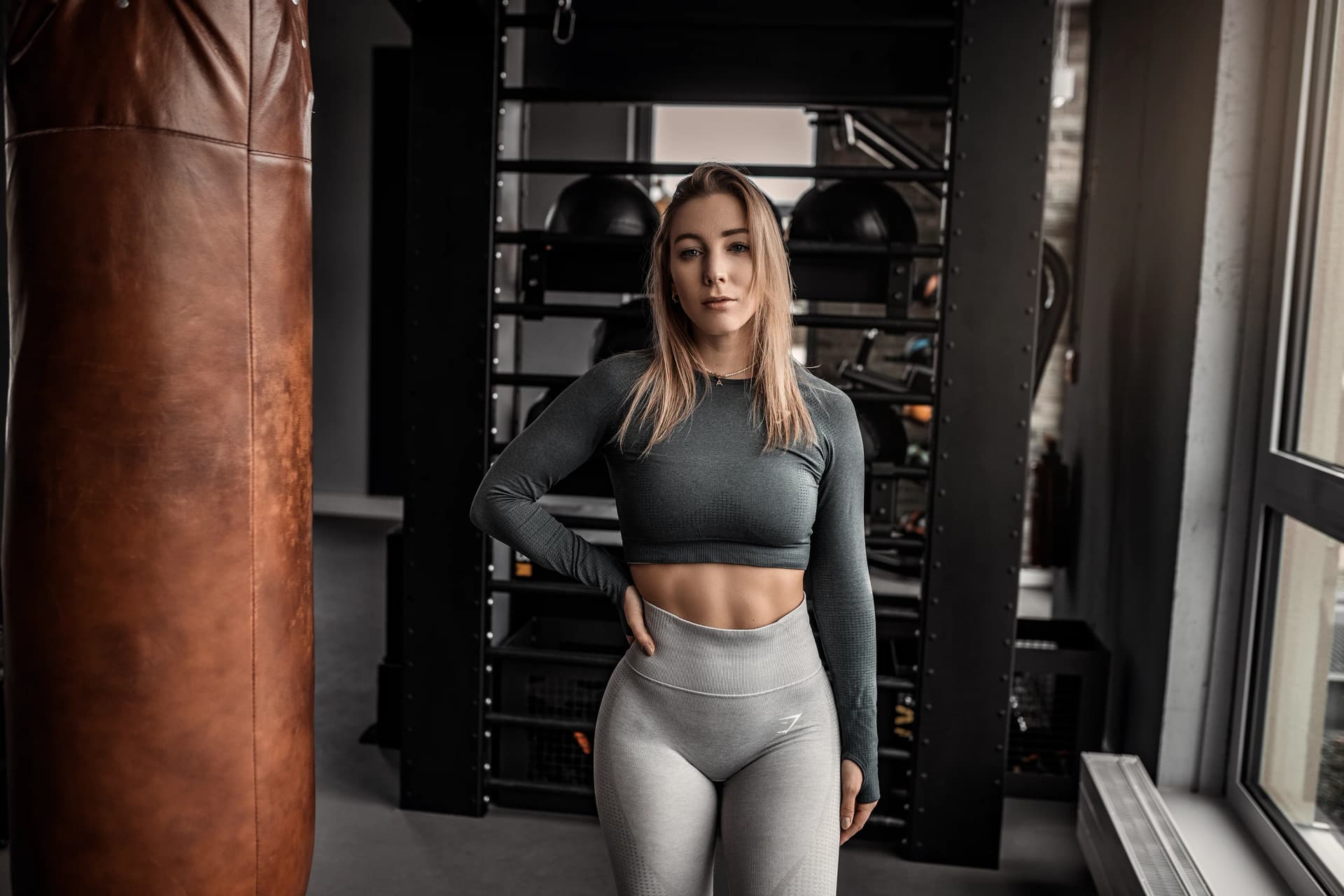 Athletin im grauen Longsleeve-Set posiert neben einem Boxsack im Studio-Gym, durchtrainierte Mitte beim Fitness-Shooting mit warmem Licht