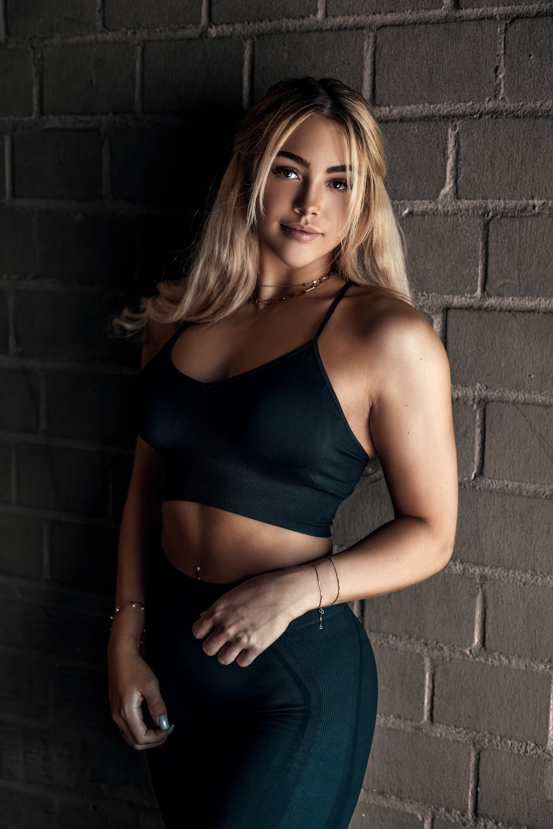 Blonde Frau im schwarzen Sport-Outfit lehnt an einer dunklen Backsteinwand, durchtrainierter Look beim Fitness-Shooting in stimmungsvollem Licht