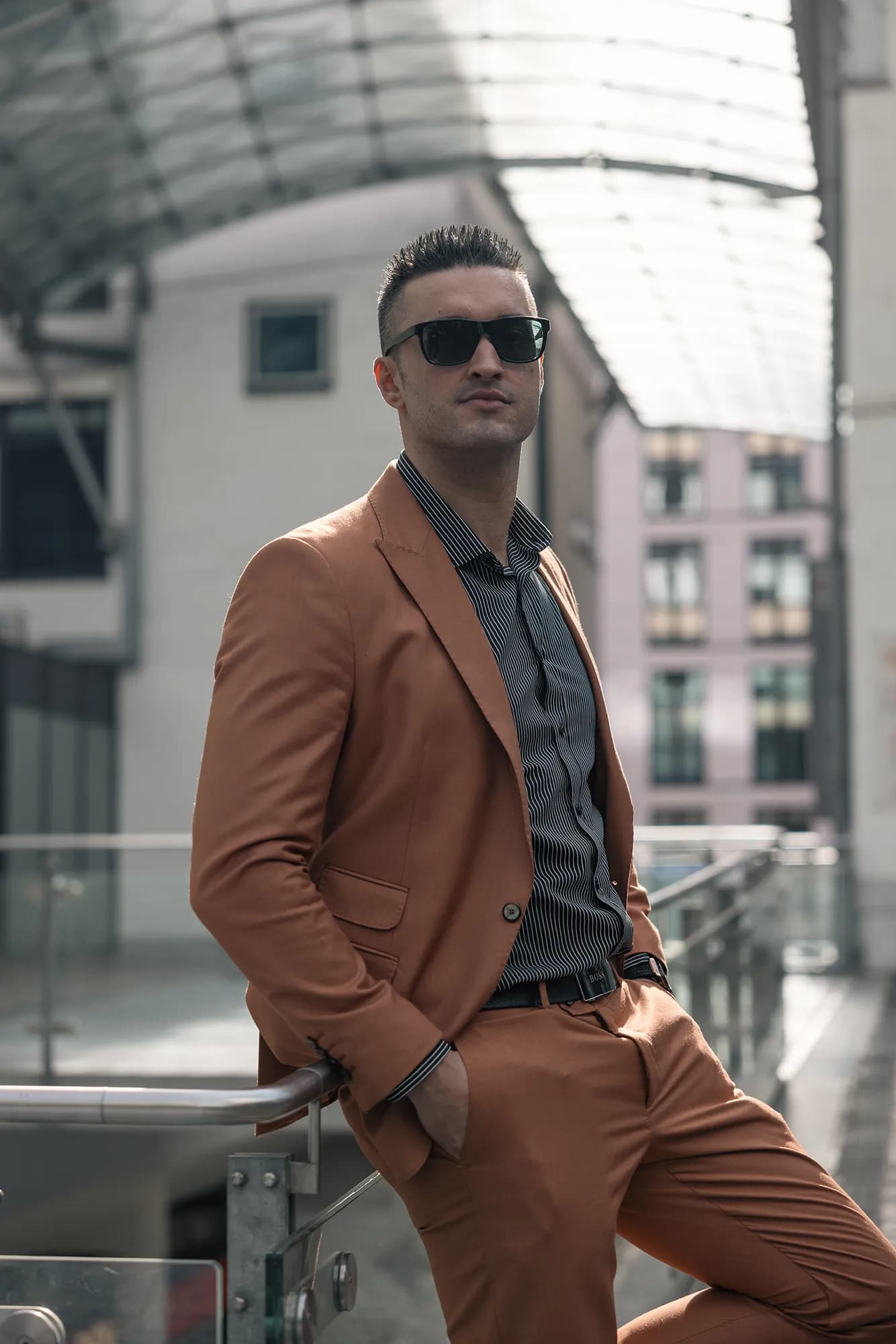 Business-Portrait eines muskulösen Mannes im cognacfarbenen Anzug und Nadelstreifenhemd mit Sonnenbrille, selbstbewusste Pose unter einer modernen Glaskuppel