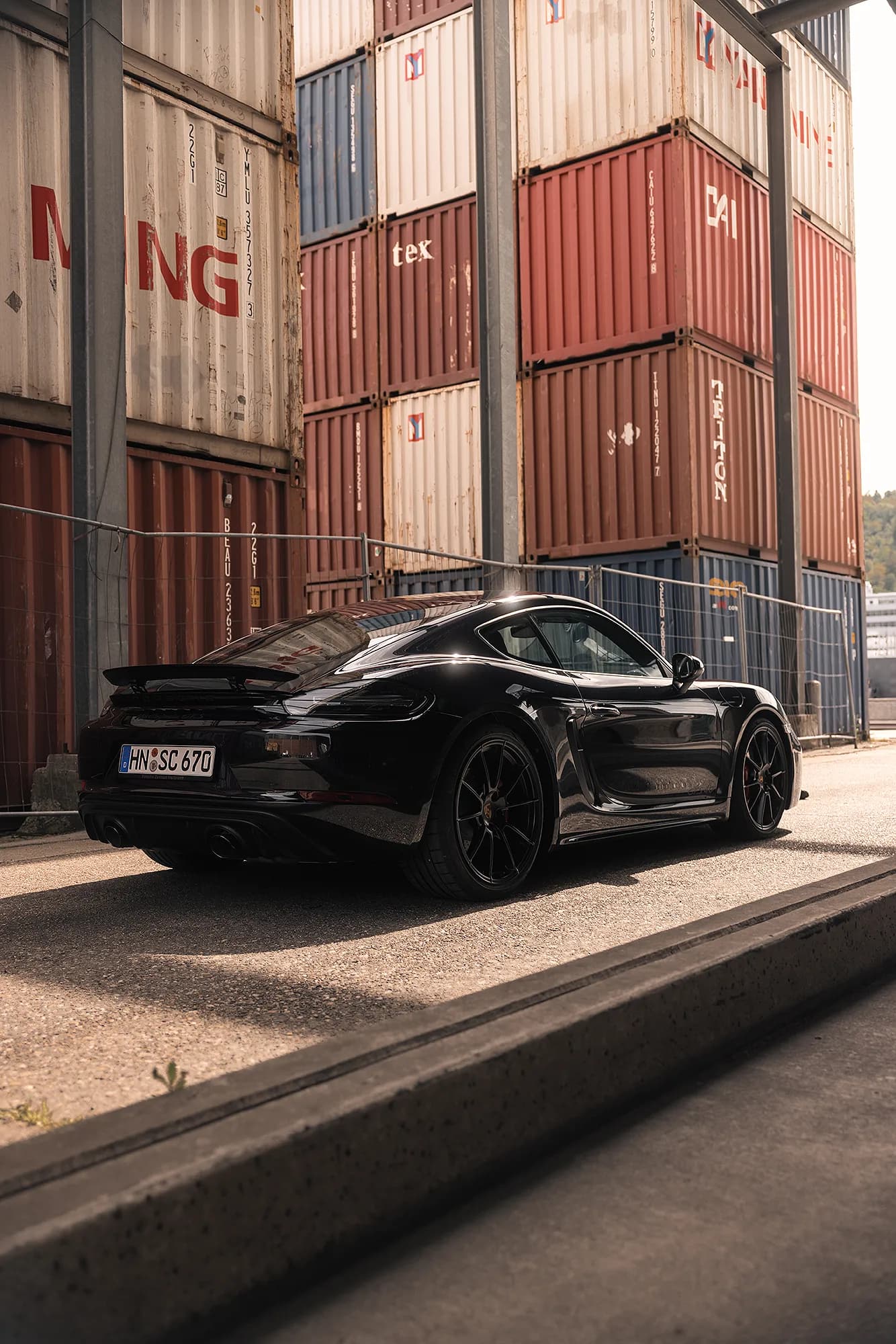 Schwarzer Sportwagen am Straßenrand vor gestapelten Seecontainern, urbane Automotive-Aufnahme im Industriegebiet