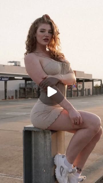 Junge Frau mit roten Locken im beigen Sommeroutfit auf einem Pfosten sitzend, urbanes Editorial im Abendlicht