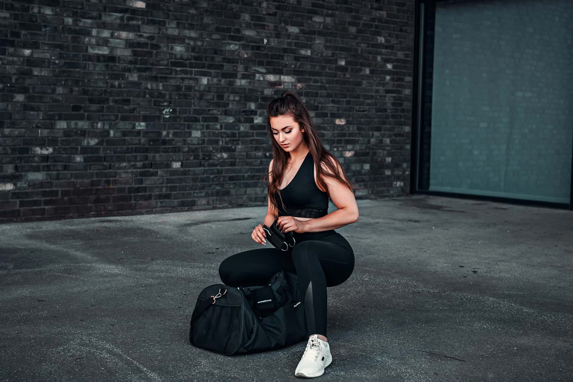 Sportlerin in schwarzem Trainingsoutfit kniet vor ihrer Sporttasche auf Asphalt, urbanes Fitness-Shooting vor rauer Backsteinkulisse