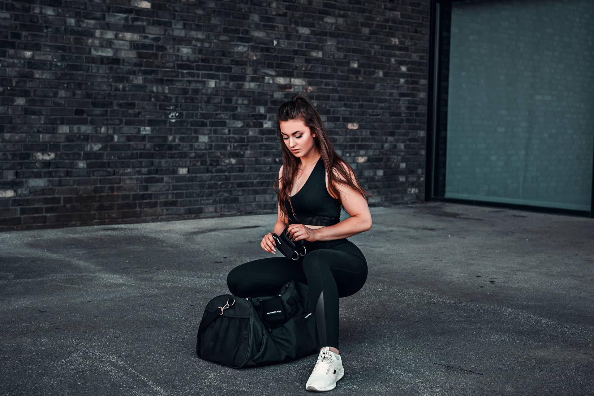 Sportlerin in schwarzem Trainingsoutfit kniet vor ihrer Sporttasche auf Asphalt, urbanes Fitness-Shooting vor rauer Backsteinkulisse