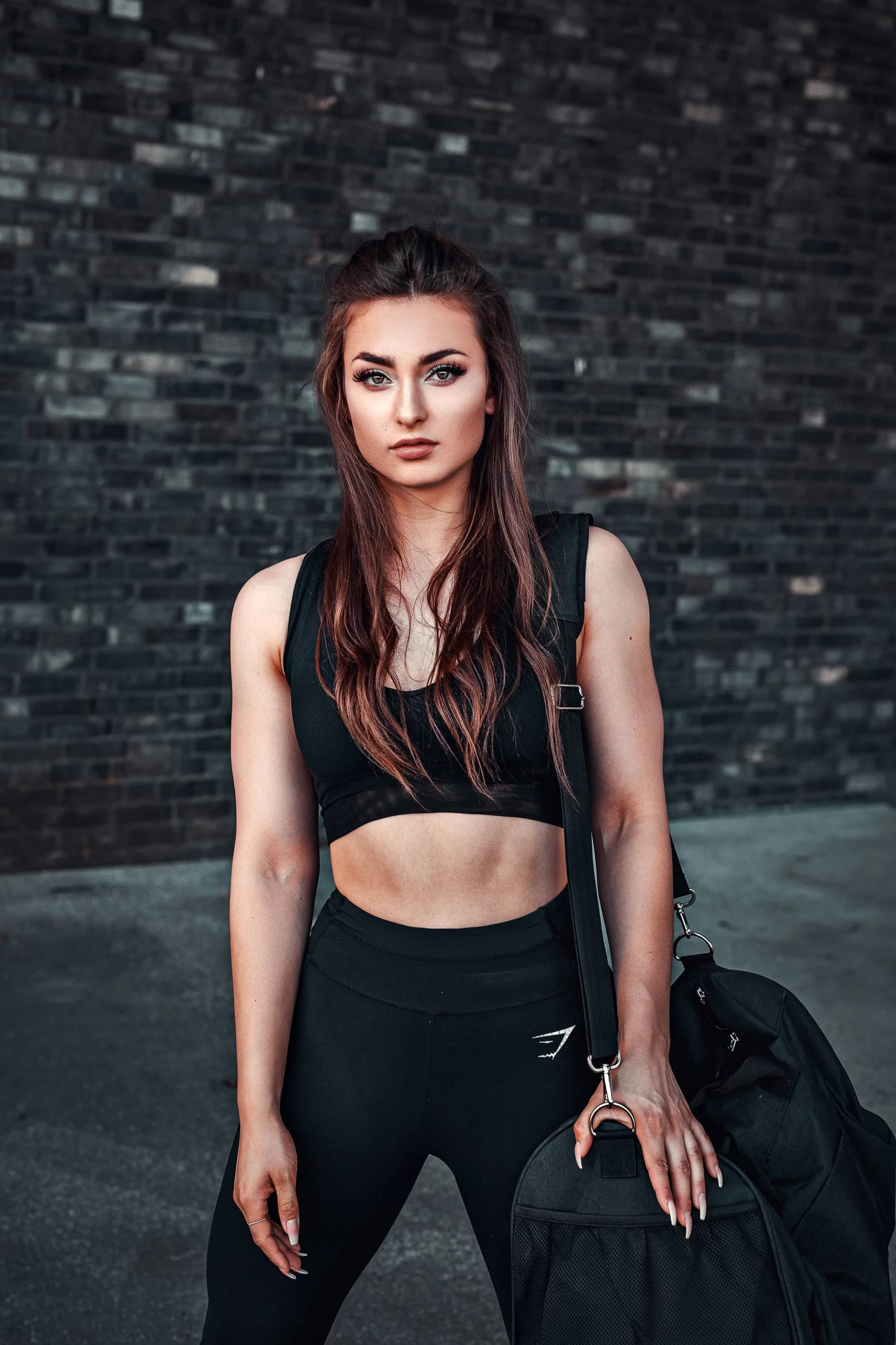 Sportliche Frau im schwarzen Trainingsoutfit mit Sporttasche vor dunkler Klinkerwand, urbanes Fitness-Shooting mit kühler, kontrastreicher Stimmung