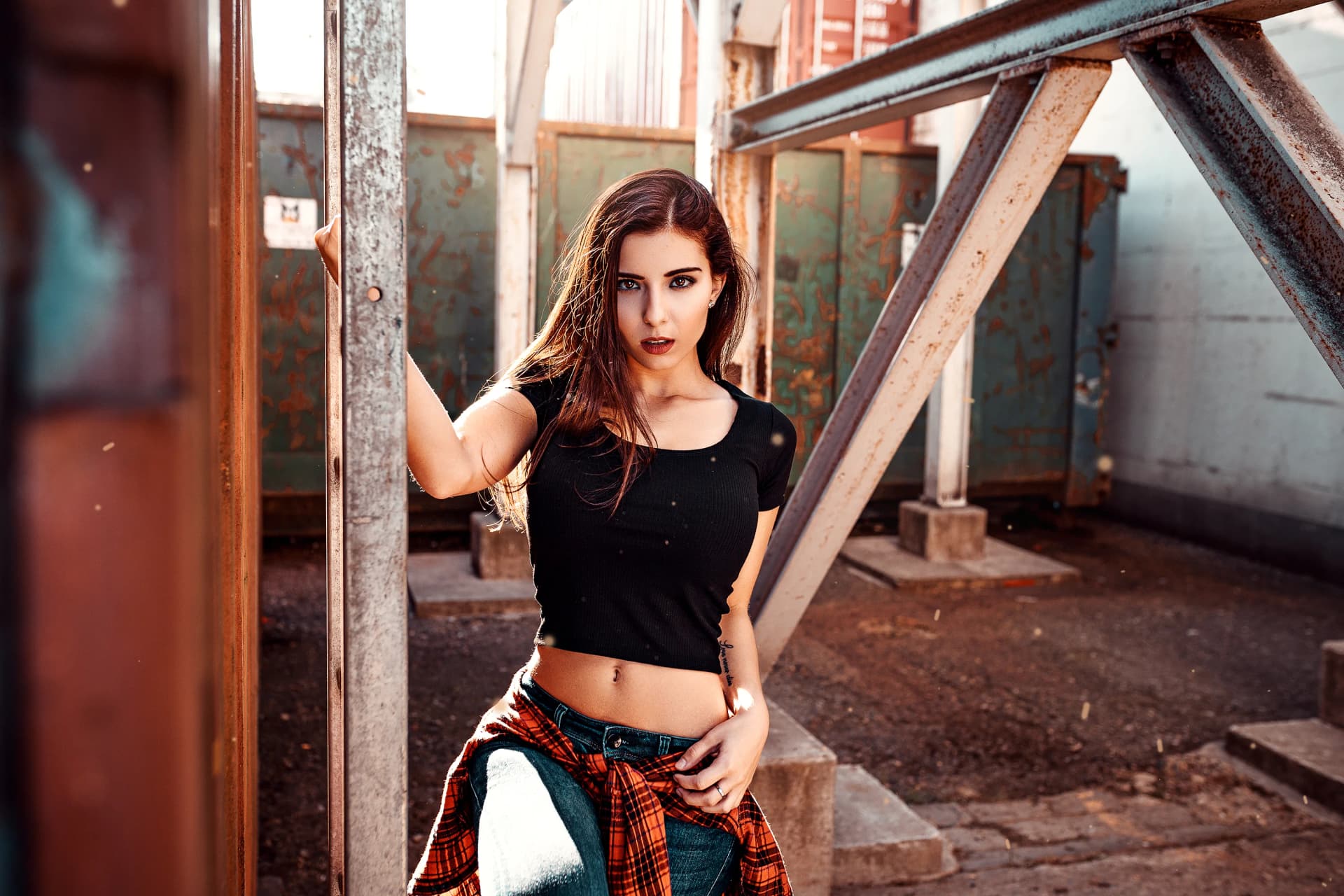 Frau in schwarzem Shirt und Jeans mit umgebundenem Karohemd in einer Industriekulisse mit rostigen Stahltraegern, dynamisches Portraitshooting im warmen Gegenlicht