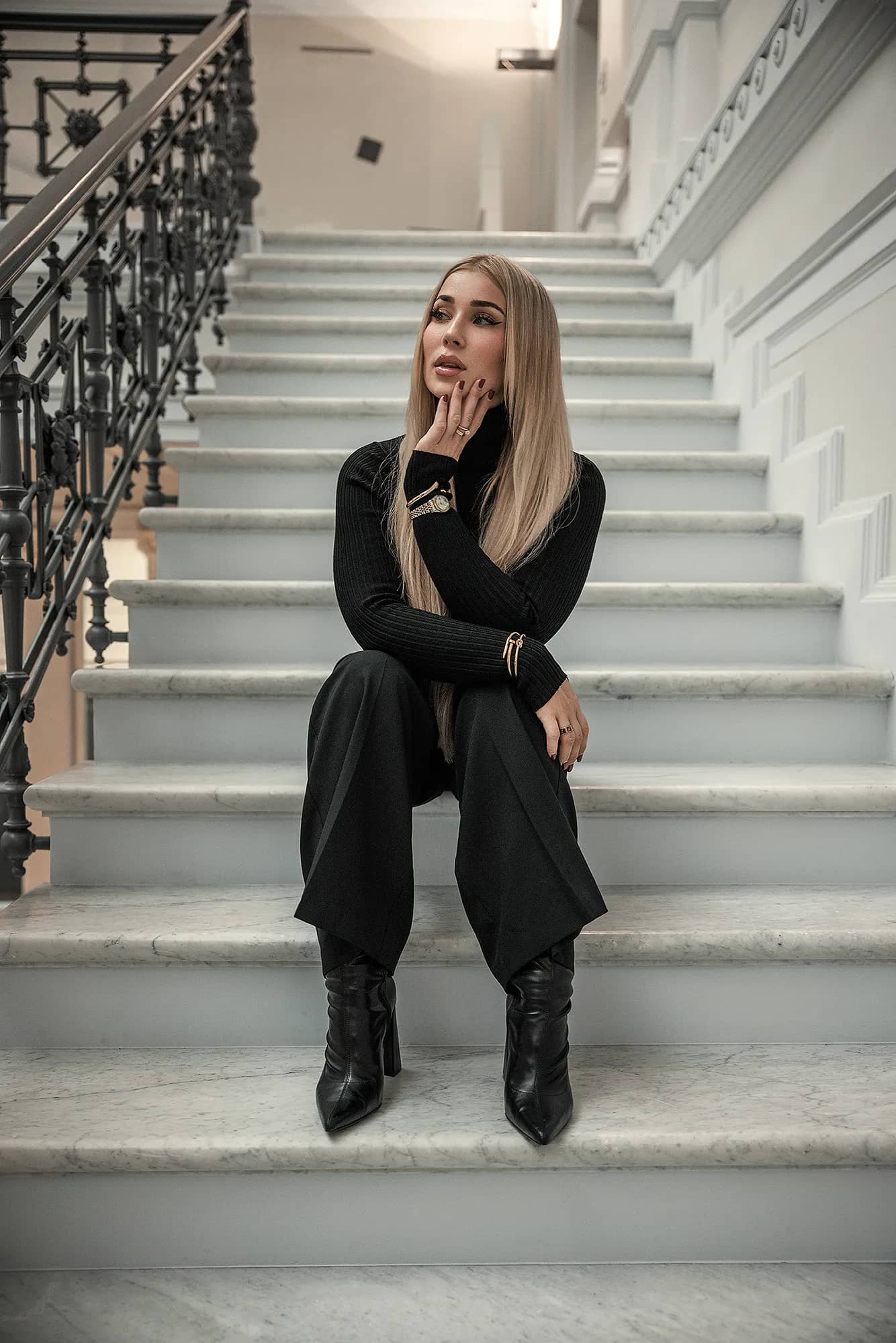 Blonde Frau in schwarzem Rollkragen und weiter Hose sitzt nachdenklich auf einer hellen Marmortreppe, edles Fashion-Shooting mit kuehlem Licht